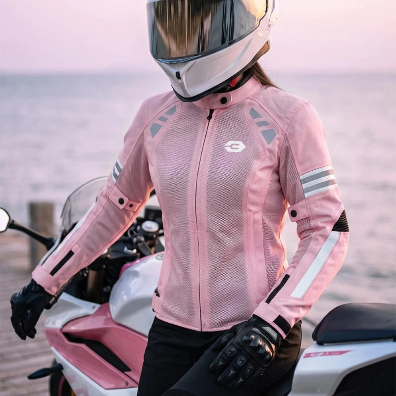 A person wearing a pink motorcycle jacket, black gloves, and a white full-face helmet with cat-ear-like fluffy details stands beside a pink and white motorcycle near the water at sunset.