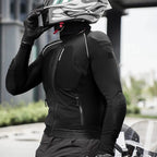 A person wearing a black full-face motorcycle riding suit jacket and matching gloves, holding a white and gray flame-patterned motorcycle helmet in an outdoor setting.