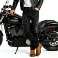 A person wearing black leather jacket, black motorcycle jeans with contrast stitching, and brown boots, posing next to a black Harley-Davidson motorcycle on a white studio background.