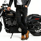 A person wearing black leather jacket, black motorcycle jeans with contrast stitching, and brown boots, posing next to a black Harley-Davidson motorcycle on a white studio background.