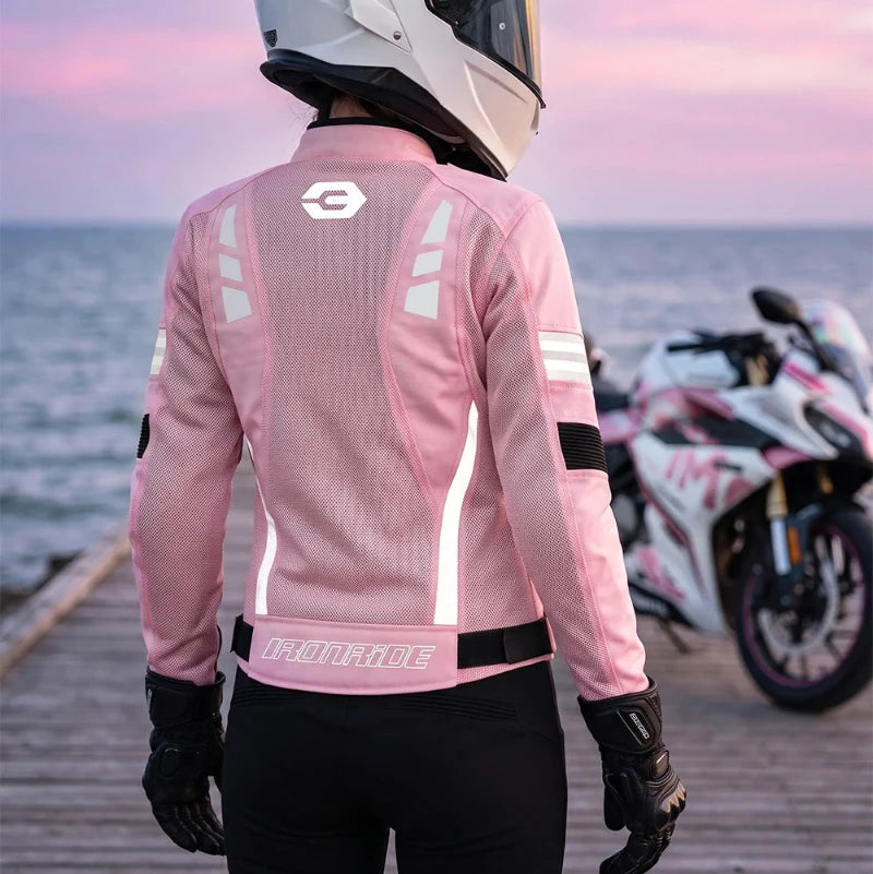 A person wearing a pink motorcycle jacket, white helmet, and black gloves stands on a wooden dock by the sea at sunset, with a white and pink motorcycle parked in the background.