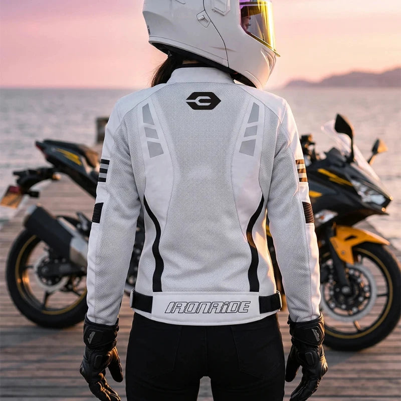 A person wearing a white full-face motorcycle helmet, a white IronRide jacket, and black pants stands on a wooden dock with two motorcycles in the background at sunset.