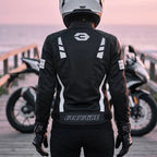 A person wearing a black and white motorcycle jacket, black pants, and a white full-face helmet stands on a wooden boardwalk with motorcycles in the background at sunset. The jacket has white graphic stripes and a visible IronRide logo on the back.