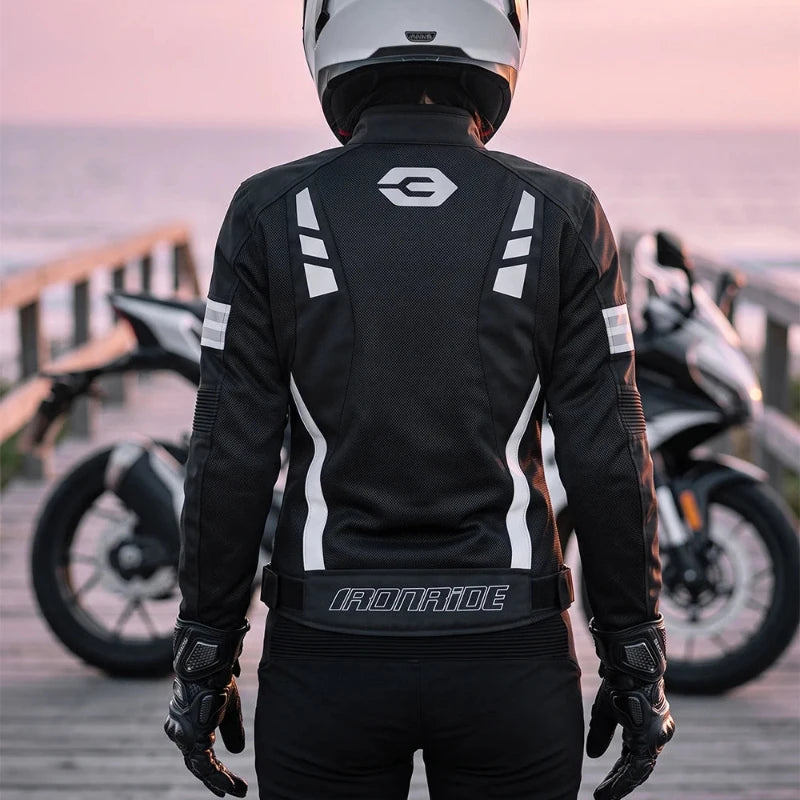A person wearing a black and white motorcycle jacket, black pants, and a white full-face helmet stands on a wooden boardwalk with motorcycles in the background at sunset. The jacket has white graphic stripes and a visible IronRide logo on the back.