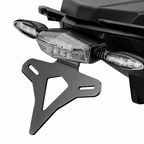 A black metal motorcycle license plate frame shown mounted against the rear body of a black motorcycle, with the license light and side indicators visible, on a plain white background.