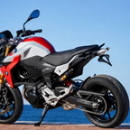 A BMW motorcycle, specifically a Motosport model, shown from a rear three-quarter angle on a paved path with the ocean and blue sky in the background. The bike features a white and red body with black seat, black wheels, and gold suspension forks.