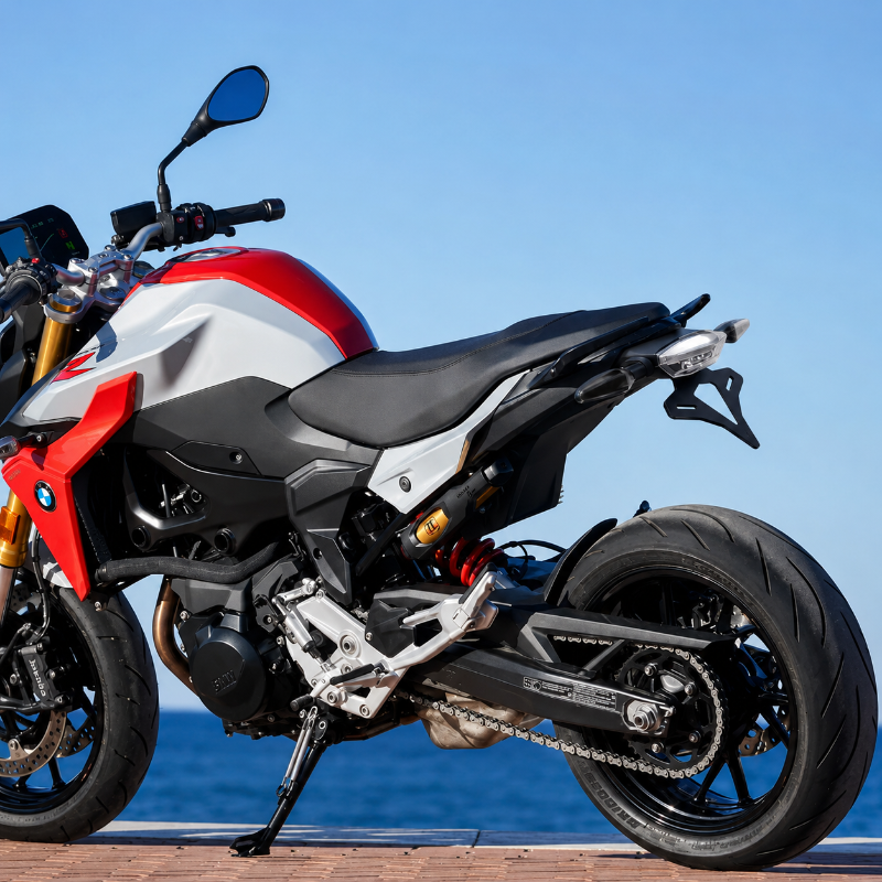 A BMW motorcycle, specifically a Motosport model, shown from a rear three-quarter angle on a paved path with the ocean and blue sky in the background. The bike features a white and red body with black seat, black wheels, and gold suspension forks.