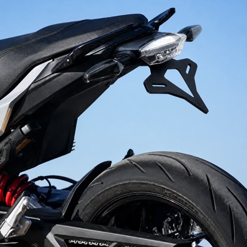Close-up of a black motorcycle rear section with a black license plate frame, rear light, tire, and red rear spring against a clear blue sky.
