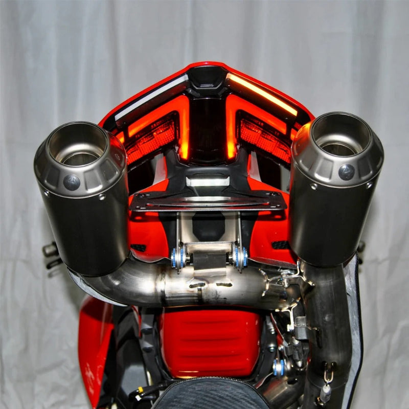A close-up of a motorcycle's exhaust system featuring two large, cylindrical black mufflers with polished stainless steel pipes connecting them, set against a red and black motorcycle body and a light gray fabric backdrop.