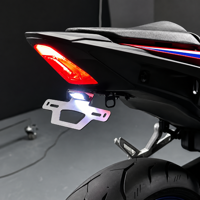 A close-up studio shot of a motorcycle rear section showing a white LED license plate light mounted below the taillight, with the license plate illuminated. The motorcycle has a black body with red and blue accent stripes.