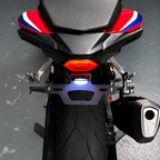 Close-up rear view of a motorcycle showing a black metal license plate frame with an integrated blue LED light, mounted above a black rear tire on a gray floor.