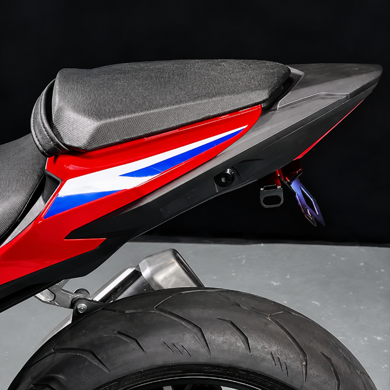 Close-up rear view of a sport motorcycle showing the black seat, red body panel with blue and white graphic stripes, black rear fender, exhaust, and tire against a dark studio background.