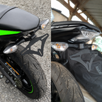 A black metal motorcycle rear rack bracket shown installed behind a motorcycle in two close-up views, with gravel pavement on the left and a covered vehicle under a shelter on the right.