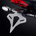 A close-up of a silver metal motorcycle license plate frame mounted on a black handlebar, with a red taillight visible in the background against a dark studio setting.