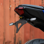Close-up of a black motorcycle license plate frame and rear light assembly mounted on a black motorcycle body, with a red-orange corrugated metal wall in the background.