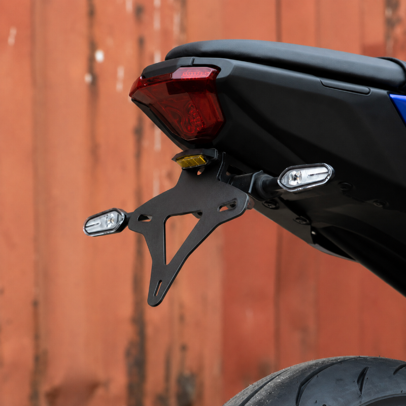 Close-up of a black motorcycle license plate frame and rear light assembly mounted on a black motorcycle body, with a red-orange corrugated metal wall in the background.