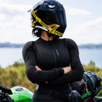 A woman wearing a black full-face motorcycle helmet with yellow caution-style tape around the visor, a black long-sleeve riding jacket, and black pants stands with arms crossed in front of a green motorcycle outdoors near water.