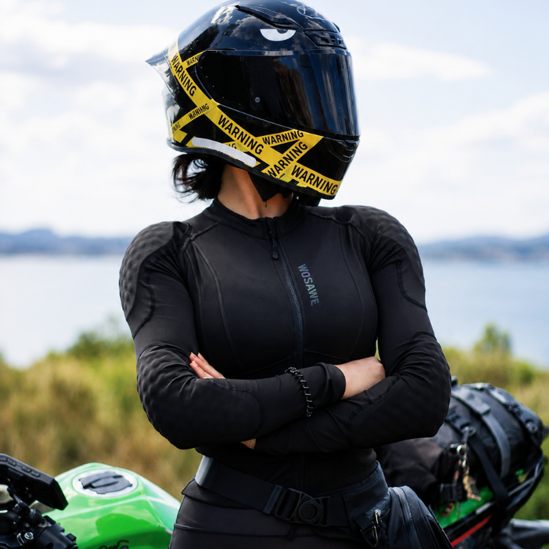 A woman wearing a black full-face motorcycle helmet with yellow caution-style tape around the visor, a black long-sleeve riding jacket, and black pants stands with arms crossed in front of a green motorcycle outdoors near water.