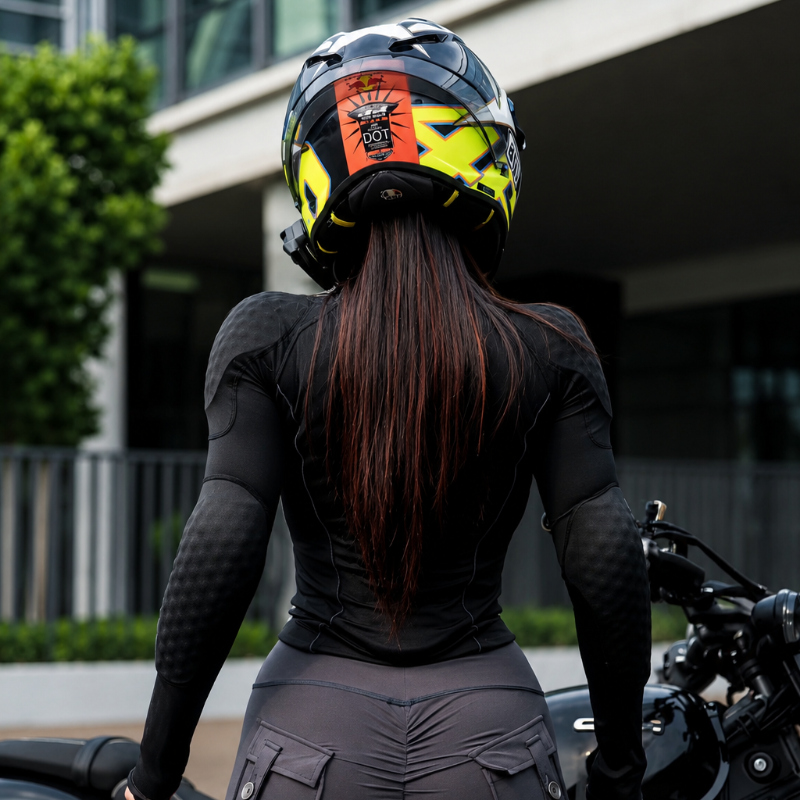 A woman seen from behind wearing a black motorcycle riding jacket and matching dark gray pants, seated on a motorcycle outdoors. She is wearing a black full-face helmet with bright yellow and orange graphics.