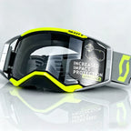 motocross goggles with a black and neon yellow frame, a clear impact-resistant lens, and a gray strap with a neon yellow logo, shown on a reflective light gray background.