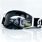 motocross goggles with a black and white frame, dark tinted lens, and a black and white strap, shown on a reflective white background.