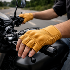 A close-up of a person riding a motorcycle, wearing tan leather fingerless gloves on both hands. The motorcycle handlebars and dashboard are visible in the foreground, with a blurred road and greenery in the background.
