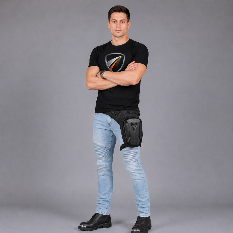 A man wearing a black t-shirt, light blue jeans, and black boots stands with arms crossed, wearing a black fanny pack with a shield logo on his hip against a gray studio background.