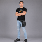 A man wearing a black t-shirt, light blue jeans, and black boots stands with arms crossed, wearing a black fanny pack with a shield logo on his hip against a gray studio background.