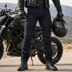 A person wearing black biker pants, a black leather jacket, black gloves, and black boots stands beside a black motorcycle on an outdoor road, holding a black helmet.