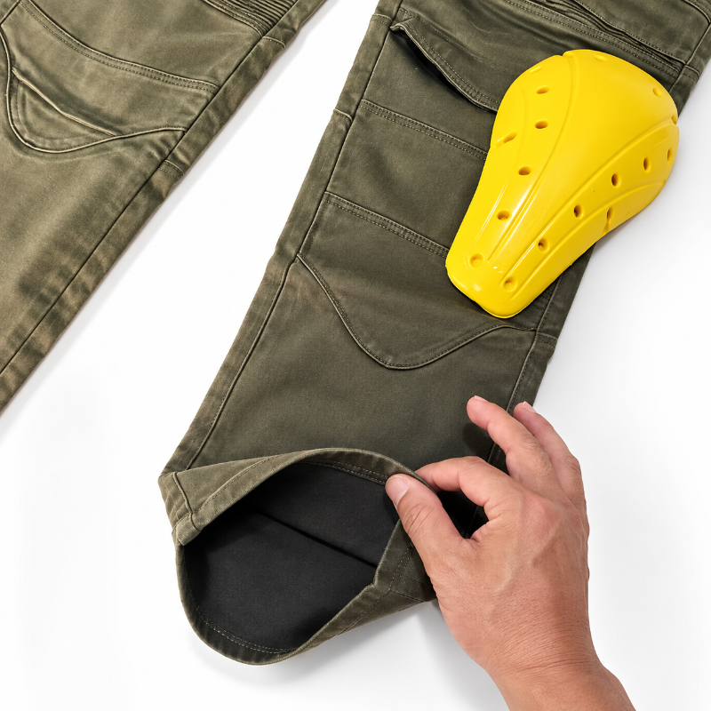 Olive green tactical pants with a hand pulling up the hem to reveal a black interior lining, featuring a yellow protective pad in the cargo pocket, on a white background.
