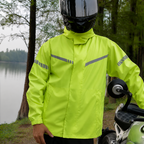 A person wearing a bright neon yellow-green rain jacket with reflective gray stripes stands outdoors beside a motorcycle, with trees and water in the background.