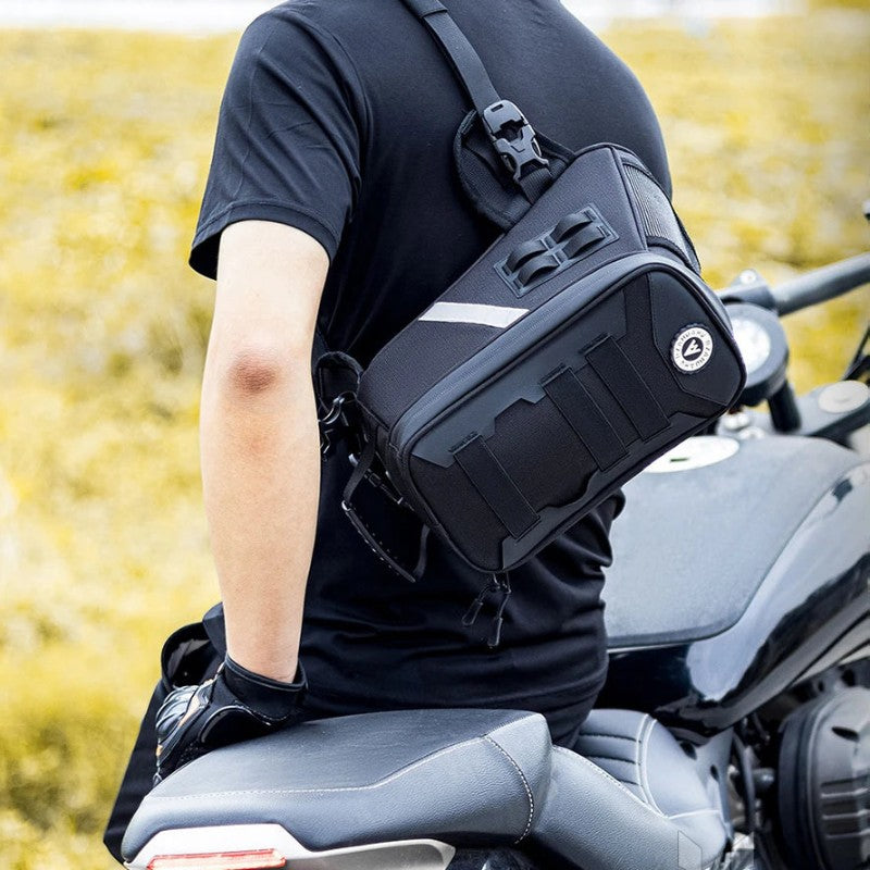 A black motorcycle side bag with a silver reflective stripe and a circular logo badge, worn crossbody on a person sitting on a motorcycle in an outdoor field.