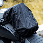 A black motorcycle tank cover with a crinkled, waterproof texture, shown fitted on a motorcycle tank in an outdoor setting with a blurred yellow background.