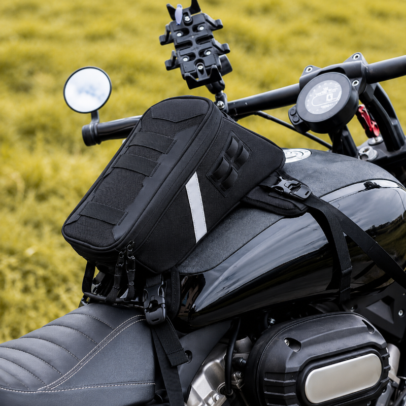 A black motorcycle tank bag with a white reflective stripe, mounted on the side of a black motorcycle with a blurred yellow-green grass background.