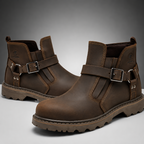 A pair of dark brown leather ankle boots with buckle straps, shown on a neutral gray gradient background.