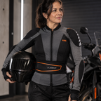 A woman wearing a gray and black motorcycle jacket with a black waist protector and holding a black helmet, standing in front of a motorcycle in a garage setting.